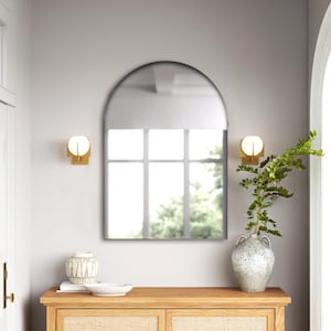 May include: An arched wall mirror with a thin black frame. The mirror reflects a window with a grid pattern. It is mounted on a light gray wall between two gold-toned wall sconces with spherical white glass shades, positioned above a wooden console.