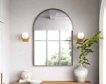 May include: An arched wall mirror with a thin black frame. The mirror reflects a window with a grid pattern. It is mounted on a light gray wall between two gold-toned wall sconces with spherical white glass shades, positioned above a wooden console.