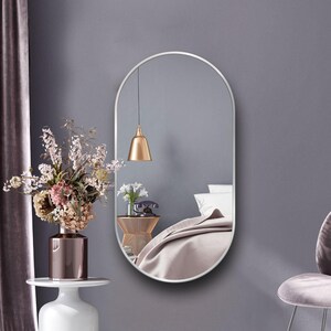Puede incluir: Un espejo ovalado plateado con un marco fino cuelga de una pared gris claro. El espejo refleja un dormitorio con una cama, una l&aacute;mpara y una mesita de noche.