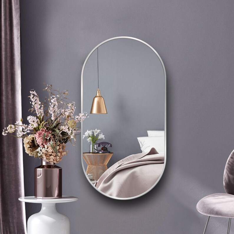 Puede incluir: Un espejo ovalado plateado con un marco fino cuelga de una pared gris claro. El espejo refleja un dormitorio con una cama, una l&aacute;mpara y una mesita de noche.