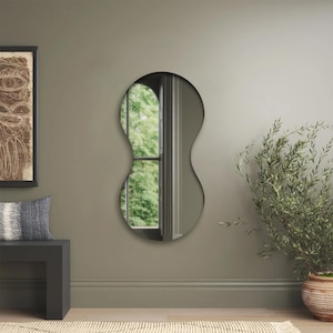 May include: A wall-mounted mirror with a distinct peanut-shaped silhouette, featuring a smooth, continuous frame that curves inward at the center. The reflective glass surface shows a clear view of an outdoor window scene with green foliage.