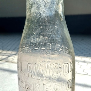 May include: A clear glass milk bottle with embossed lettering. The bottle reads "HERFIELD FARM" and "LAWSON BECKER CO. NEW YORK". The bottle has a rounded base and a narrow neck. The glass has a slightly frosted appearance.