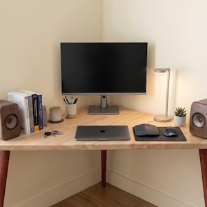 Può includere: Una scrivania angolare triangolare in legno con tre gambe affusolate color rosso scuro. Sulla scrivania ci sono un monitor per computer, un portatile chiuso, due altoparlanti in legno, una lampada, una piccola pianta grassa, una tazza, un portapenne e una pila di libri.