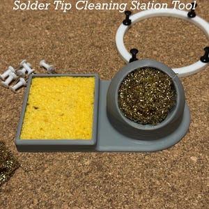 Pode incluir: Ferramenta de limpeza de ponta de solda com base cinza, esponja amarela e escova de arame de latão. A ferramenta está sobre uma superfície de cortiça.