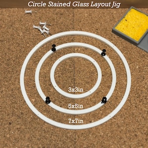 Peut inclure: Gabarit circulaire en plastique blanc avec trois cercles concentriques pour des projets de vitrail. Les cercles sont étiquetés 3x3in, 5x5in et 7x7in. Quatre punaises noires maintiennent le gabarit en place sur un tableau en liège.