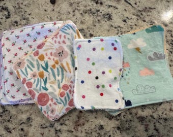 May include: A collection of soft, square baby washcloths. The cloths feature various patterns, including floral designs in pink and yellow, colorful polka dots, and a cloud and rain design in blue and pink. The edges are finished with a contrasting trim.