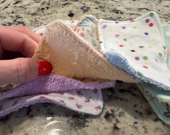 May include: A stack of colorful, reusable face cloths. The cloths have various patterns, including polka dots, floral prints, and solid colors. The edges are trimmed with different colored fabrics. The cloths are made of soft, absorbent material.