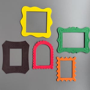 Puede incluir: Cinco marcos de fotos coloridos en amarillo, verde, naranja, rojo y marrón están dispuestos sobre un fondo gris. Los marcos tienen varias formas, incluyendo diseños cuadrados y arqueados, y están hechos de papel grueso.