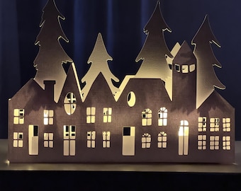 May include: A papercraft village scene with a lighted interior. The village is made of gray paper and features several houses, a church, and trees. The houses have windows and doors cut out, and the church has a steeple. The trees are silhouetted against the background.