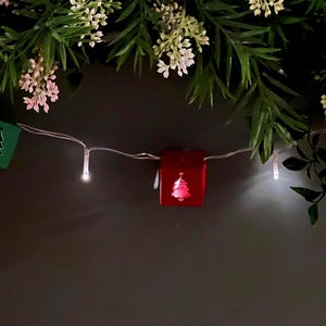 May include: A festive Christmas decoration featuring a string of lights with small, illuminated boxes. The boxes are red, green, and gold, each with a cutout or design of a Christmas tree. Greenery and white flowers are arranged above.