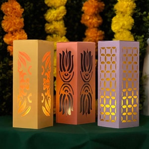 May include: Three decorative paper lanterns with intricate cut-out designs. The lanterns are in shades of yellow, orange, and purple, and are illuminated from within.