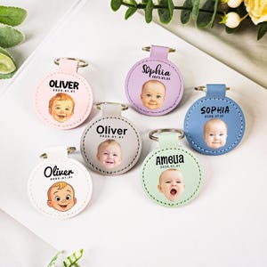 May include: A collection of personalized keychains in various pastel colors, each featuring a baby's photo and name. The keychains have a circular design with a metal ring and leather strap. Names include Oliver, Sophia, and Amelia.