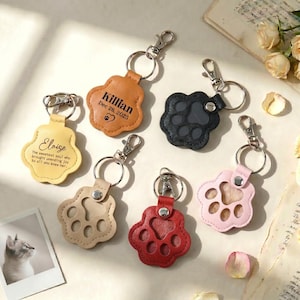 Puede incluir: Varios llaveros con forma de pata en diversos colores: beige, negro, rojo y rosa. Algunos llaveros tienen texto, como "Killian Dec 26, 2025" y "Eloise The sweetest soul who brought unending joy to all who knew her."