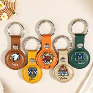 May include: Five leather AirTag keychains in shades of brown, yellow, orange, beige, and green. Each keychain has a silver ring and a circular leather tag with different designs, including team logos and text. The keychains are designed for AirTag.