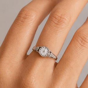 May include: A silver-toned ring featuring a miniature watch face design. The circular centre has a textured white dial with small markings, while the band displays a ribbed, metallic pattern extending from the watch face on both sides.