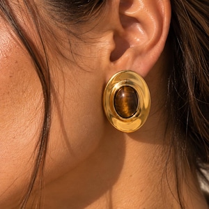 May include: An oval-shaped earring featuring a polished gold-tone metal frame surrounding a smooth, rounded tiger's eye gemstone at the center. The design showcases a warm, reflective metallic finish with the natural brown and golden banding of the stone.