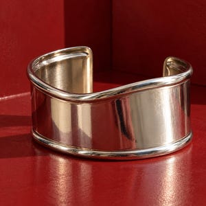 May include: A wide, polished silver-tone cuff bracelet featuring a smooth, reflective surface and a distinct undulating wave-like top edge. The open-ended design has rounded, reinforced borders along the top and bottom rims, creating a solid, metallic appearance with a high-shine finish.