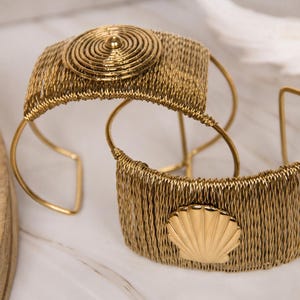 May include: Two gold-toned cuff bracelets made of fine wire mesh. One bracelet features a circular, concentric spiral design on the front, while the other bracelet displays a smooth, polished seashell emblem centered on the wire mesh surface.