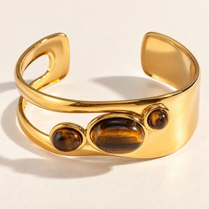 Könnte beinhalten: Ein goldfarbenes Armband in Manschettenform mit offenem Design und drei glatten, ovalen Tigerauge-Edelsteinen auf der Vorderseite. Das polierte Metallband hat eine breite, geschwungene Form mit einer Öffnung an der Rückseite.