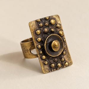 May include: A rectangular brass-toned ring featuring a textured, dark-patina surface adorned with numerous small, raised spherical studs. A prominent circular element sits in the centre, topped with a smaller dome. The band displays etched line details.