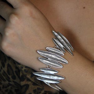 May include: Silver-toned bracelet featuring multiple elongated, leaf-shaped links. The bracelet is worn on a wrist, with a leopard print top visible in the background. The bracelet's design is modern and eye-catching.