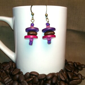 May include: A pair of earrings with a layered design. The earrings are made with purple, pink, and brown beads. The earrings are hanging from a white mug.