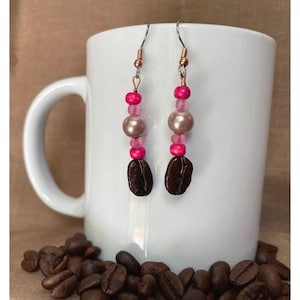 Puede incluir: Un par de pendientes colgantes con cuentas rosas y marrones. Los pendientes tienen un dije de grano de café en la parte inferior.