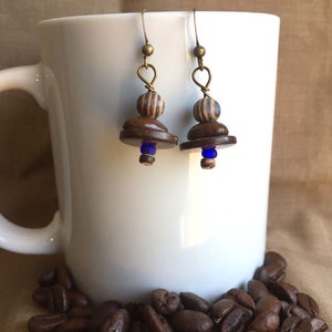 May include: A pair of brown and blue wooden earrings with a striped bead and a blue glass bead. The earrings are hanging from a white mug.