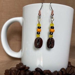 May include: Coffee bean earrings with yellow, orange, and black beads. The earrings are hanging in front of a white mug. Coffee beans are scattered at the base of the mug. The earrings have a copper-colored top.