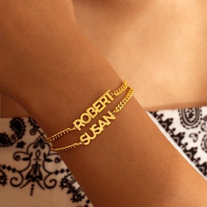 May include: Gold-colored bracelet with the names "ROBERT" and "SUSAN" in capital letters. The bracelet has a delicate chain design. The names are on separate strands, creating a layered effect. The bracelet is worn on a wrist.