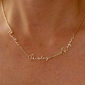 May include: A gold-colored necklace with three names: Martin, Paisley, and Regan. The names are written in a cursive script and are connected by a delicate chain. The necklace is displayed on a person's neck.