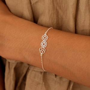 Peut inclure: Un bracelet en argent avec une chaîne délicate et trois breloques en forme de fleur. Les breloques sont en métal ajouré et présentent un motif floral.