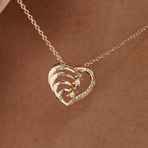 K&ouml;nnte beinhalten: Goldene herzf&ouml;rmige Halskette mit f&uuml;nf eingravierten Namen und f&uuml;nf bunten Edelsteinen. Die Halskette ist an einer Kette und wird von einer Hand gehalten.