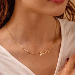 May include: A delicate gold-colored necklace with the names "Martina" and "Paisley" in cursive script. The necklace is worn around the neck, and the names are connected by a fine chain. The background is a white shirt.