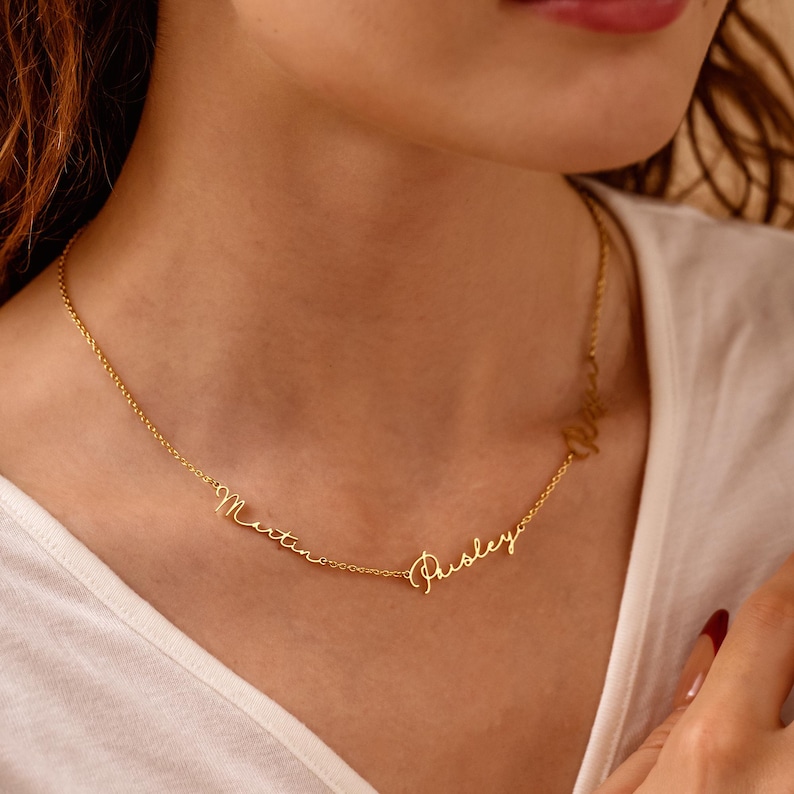 May include: A delicate gold-colored necklace with the names "Martina" and "Paisley" in cursive script. The necklace is worn around the neck, and the names are connected by a fine chain. The background is a white shirt.