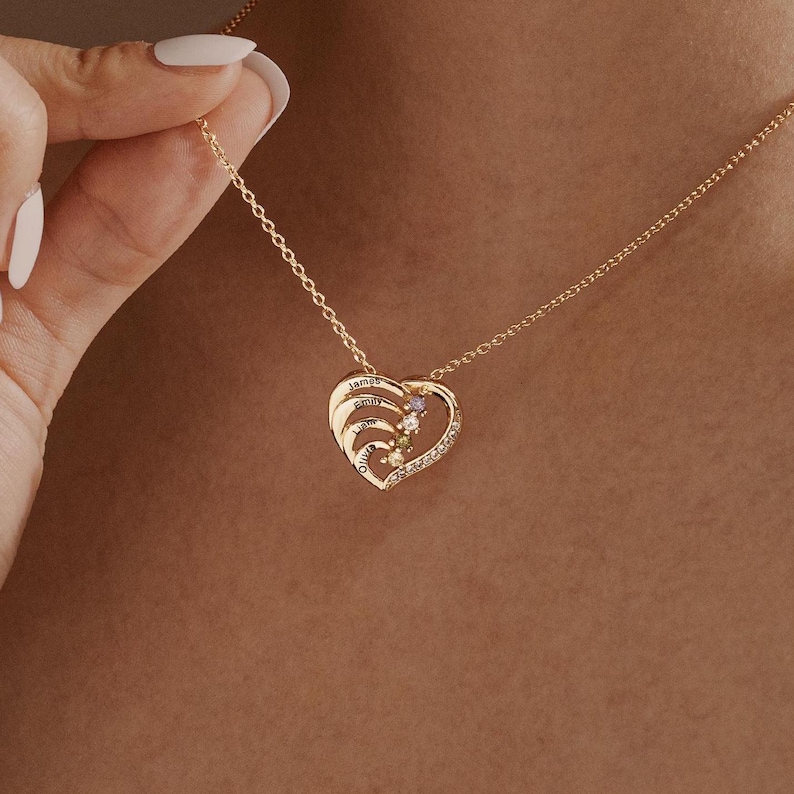 K&ouml;nnte beinhalten: Goldene herzf&ouml;rmige Halskette mit f&uuml;nf eingravierten Namen und f&uuml;nf bunten Edelsteinen. Die Halskette ist an einer Kette und wird von einer Hand gehalten.