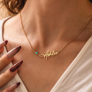 May include: A gold-colored necklace with the name "Ashlee" in a cursive font. A small turquoise stone is set to the left of the name. The necklace is worn around a person's neck.