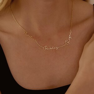May include: A delicate gold necklace with three names: Martin, Paisley, and Regan, in a cursive font. The necklace is displayed on a person's neck, showcasing its elegant design and personalized touch. The necklace is a beautiful piece of jewelry.