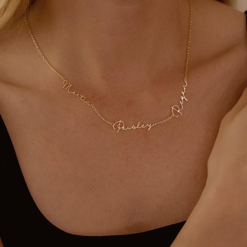 May include: A delicate gold necklace with three names: Martin, Paisley, and Regan, in a cursive font. The necklace is displayed on a person's neck, showcasing its elegant design and personalized touch. The necklace is a beautiful piece of jewelry.