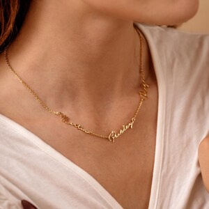 May include: Gold-colored necklace with three names in a cursive font. The names are connected by a delicate chain. The necklace is worn on a person wearing a white shirt.