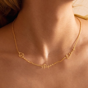 May include: A gold-colored necklace with three cursive name pendants: "Diana", "Piper", and "Elvis". The delicate chain is visible against the skin, showcasing the personalized jewelry.