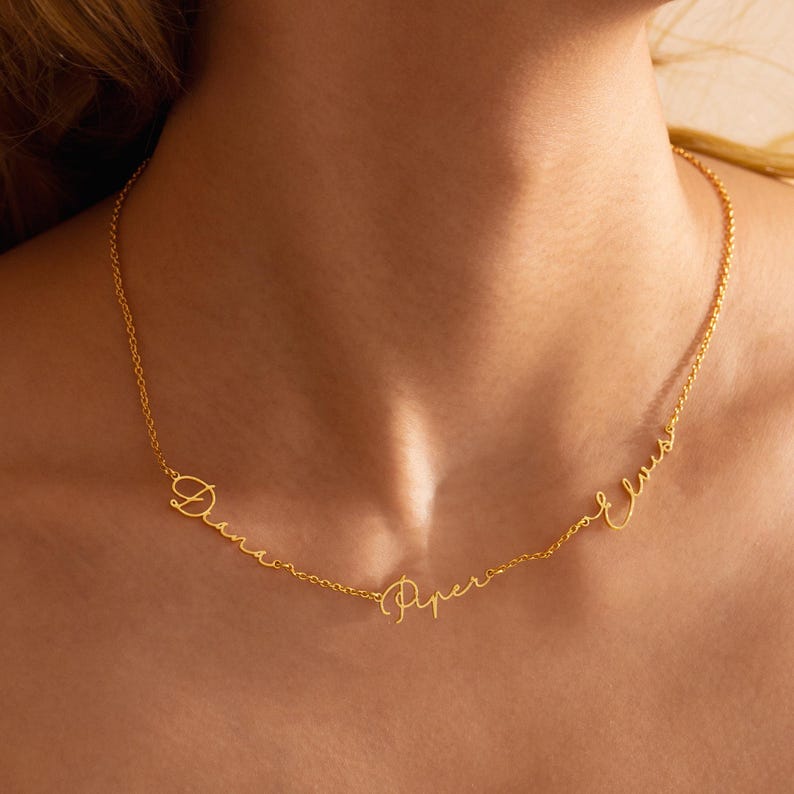 May include: A gold-colored necklace with three cursive name pendants: "Diana", "Piper", and "Elvis". The delicate chain is visible against the skin, showcasing the personalized jewelry.