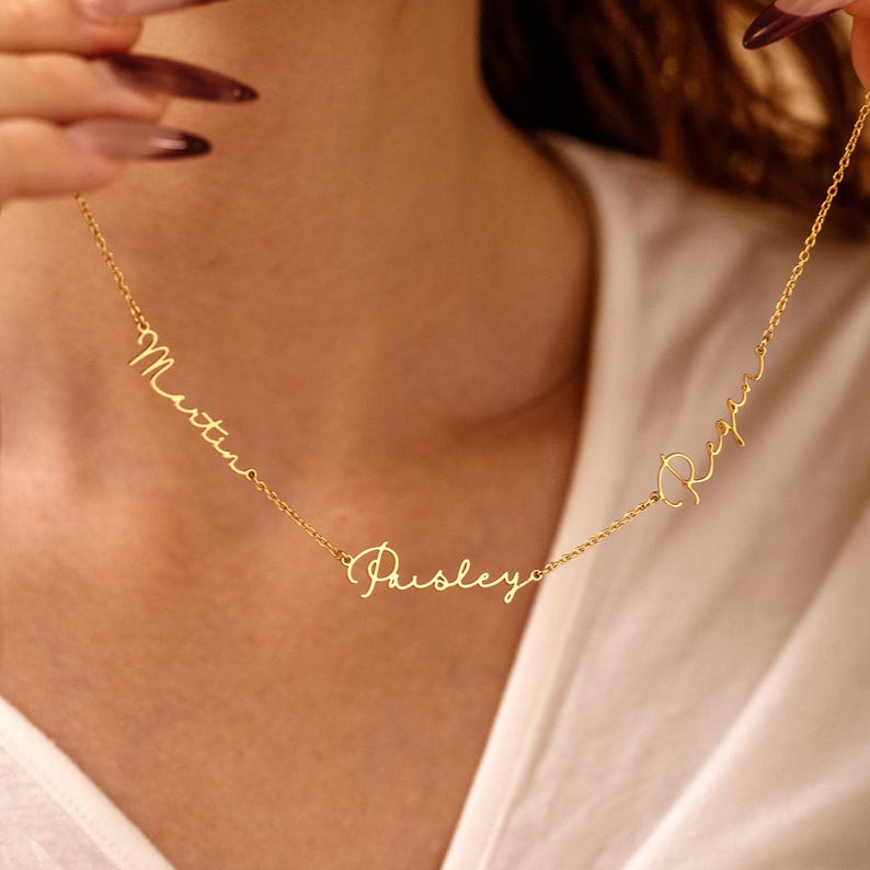 May include: A delicate gold-colored necklace featuring three names: Martina, Paisley, and Regan, written in a cursive font. The necklace is displayed against a neutral background, highlighting the jewelry's design and craftsmanship.