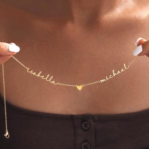 Può includere: Una collana color oro con una catenina sottile che riporta i nomi Isabella e Michael scritti in corsivo. Un piccolo ciondolo a forma di cuore pieno color oro è posizionato centralmente tra i due nomi lungo la catena.