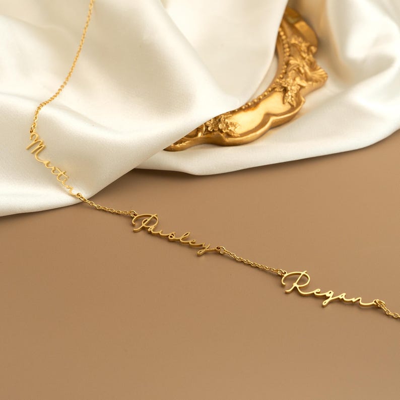 May include: A gold-colored necklace with three name pendants: "Martin", "Paisley", and "Regan". The necklace is displayed on a white satin fabric with a gold decorative element in the background. The names are written in a cursive font.
