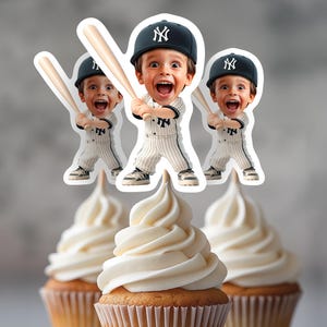 以下が含まれることがあります： 白いフロスティングが施された3つのカップケーキ。野球をテーマにしたデコレーションが施されています。デコレーションは、野球のユニフォームを着た少年とニューヨーク・ヤンキースの野球帽をかぶりバットを持った漫画のイメージです。カップケーキは明るい色の表面にあります。