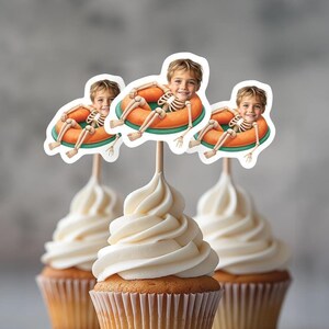 Puede incluir: Cupcakes con glaseado blanco y adornos decorativos. Los adornos representan a un niño disfrazado de esqueleto flotando en una boya naranja y verde. Los cupcakes están en moldes de papel marrón.