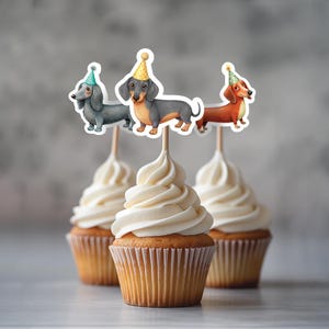 Puede incluir: Tres cupcakes con glaseado blanco y un adorno de perro salchicha en cada uno. Los dachshunds llevan sombreros de fiesta y son de diferentes colores: gris, marrón y naranja.