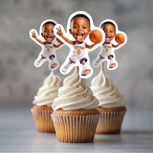 以下が含まれることがあります： 白いフロスティングが施された3つのカップケーキ。それぞれバスケットボールのユニフォームを着た子供の切り抜きがトッピングされています。子供は8番の白いジャージを着てバスケットボールを持っています。カップケーキは紙のカップに入っています。