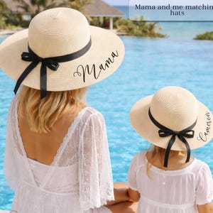 May include: Two ivory-colored sun hats with black ribbons. The larger hat has the word "Mama" printed on it, and the smaller hat has the name "Cameron". The hats are part of a matching set.